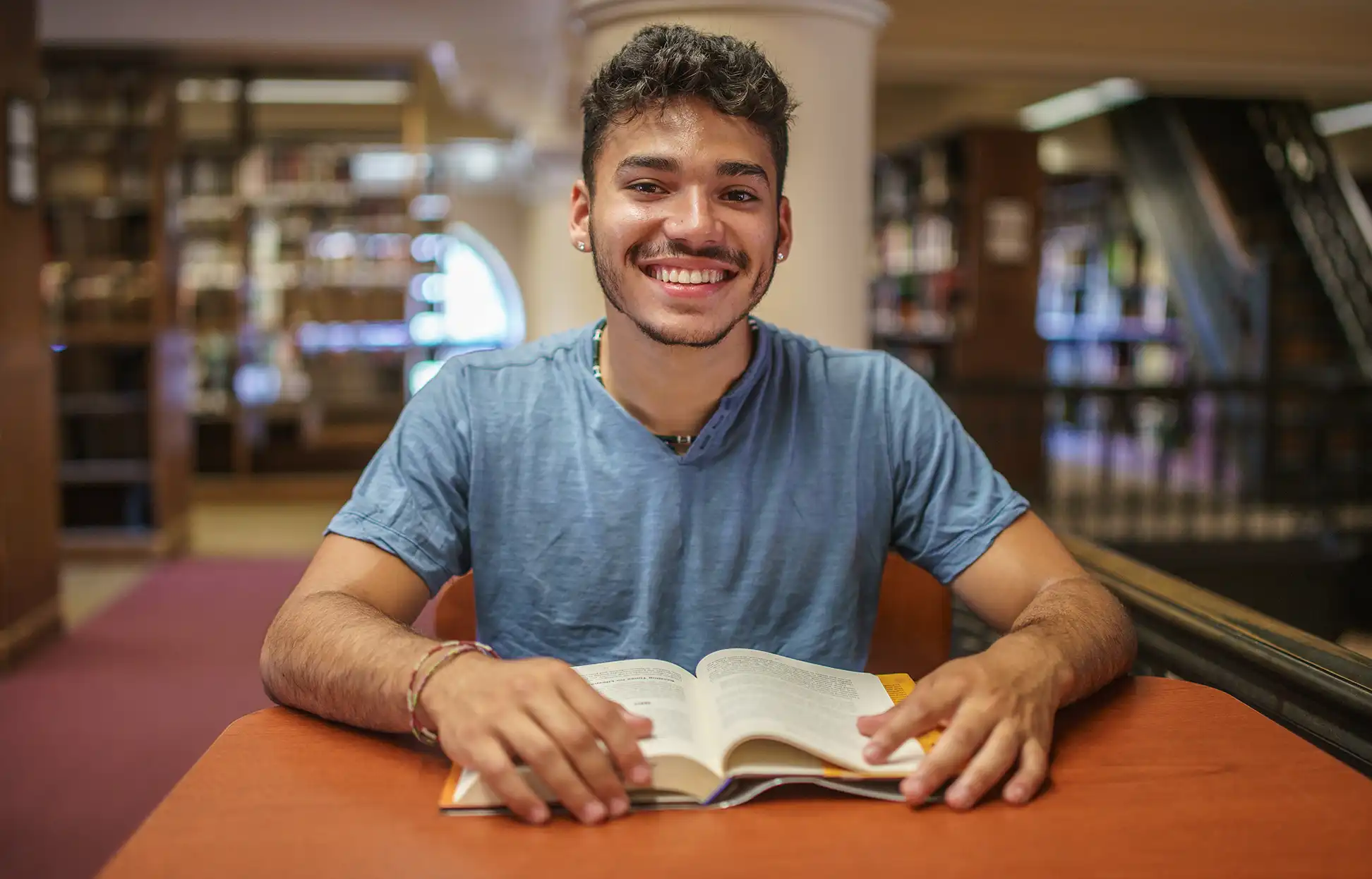 Student at a desk in a library with a book looking up and smiling